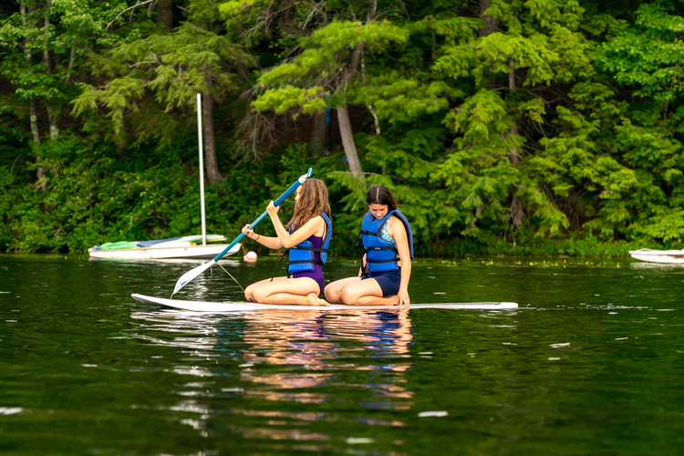 Two Session 2 Campers kneeling on a stand-up paddleboard on Hubbard Pond