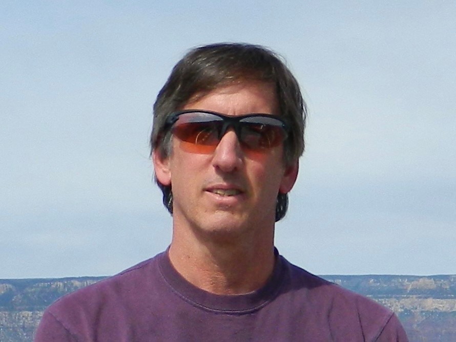 A photo of Tim Panciera from the shoulders up, wearing sunglasses and a purple t-shirt, standing in front of a low hill or cliff with blue skies in the background.