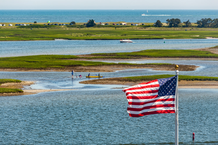 The American flag flies in the foreground over a tidal flat with kayakers in the mid-ground and the ocean in the background.