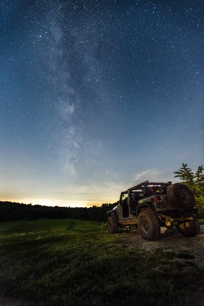 A jeep parked on a dirt road by a meadow with a star-filled sky above