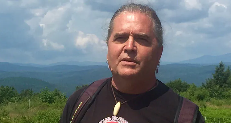 A headshot of Richard Holschuh outdoors, with green forest and mountains behind him.