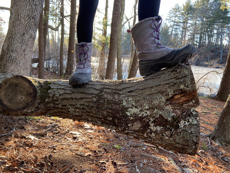 Photo from the knees-down of a child standing on a fallen log in the forest in a pair of winter boots.