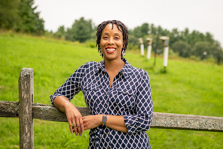 A portrait photo of Nia Keith leaning on a fence in front of a green field with bird boxes in the background. Location: Drumlin Farm Wildlife Sanctuary in Lincoln