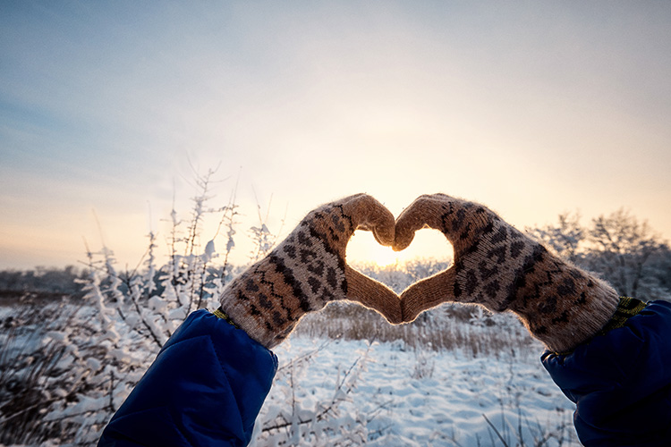 A pair of hands in knit gloves forming a heart shape against a snowy outdoor sunset