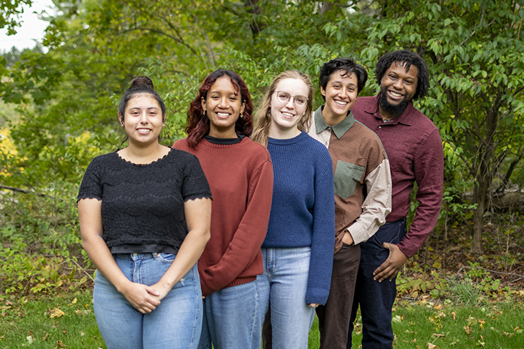 Five Environmental Fellows stand in a line smiling with green leaves in the background. 