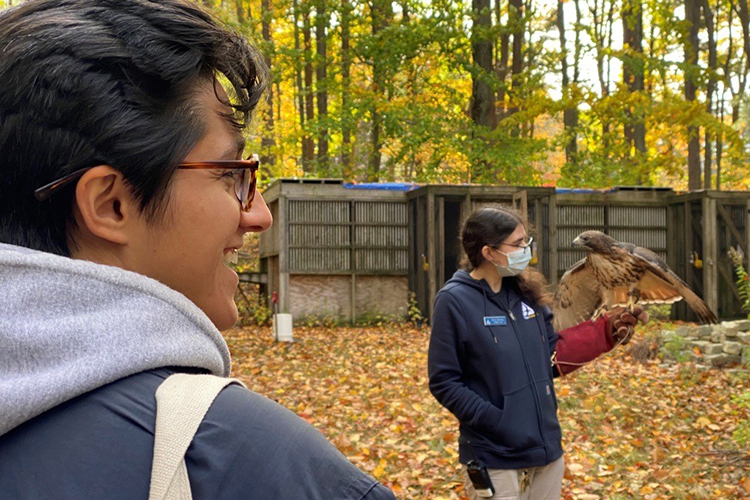 A Red-tailed hawk perches on Emily's gloved hand. In the foreground, Amara looks on, smiling. 