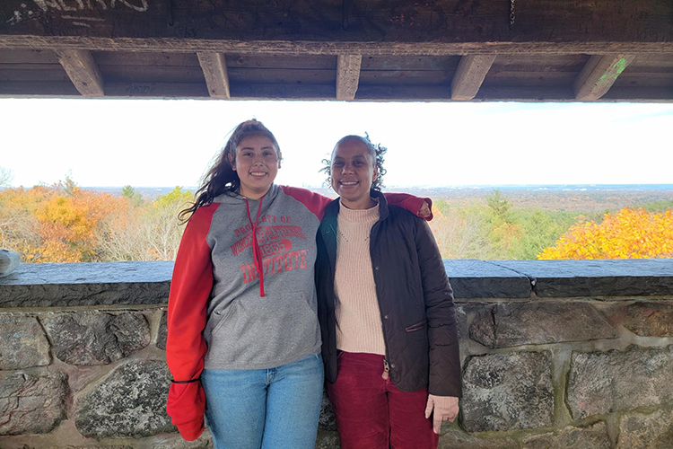 Isa and Zaskya stand together smiling and leaning on the stone wall behind them. Behind the wall is a horizon filled with fall foliage. 