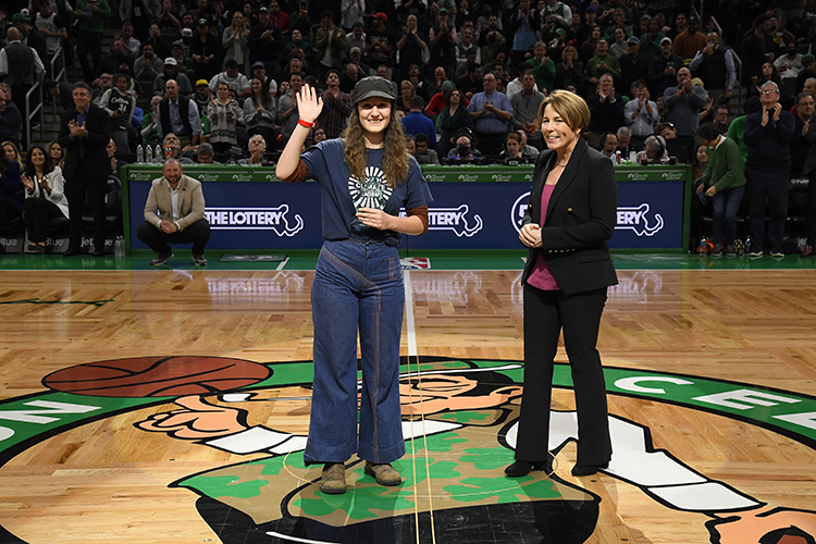 Ollie Perrault stands in the middle of a basketball court with a blue shirt and patterned jeans while waving and smiling at the camera. Standing next to her is Governor-elect Maura Healey smiling in a black blazer and black pants.