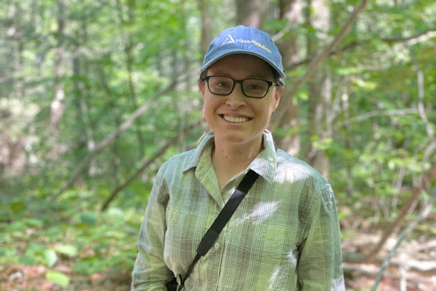 Stephanie standing in a forest, smiling with a green shirt and blue Mass Audubon hat.
