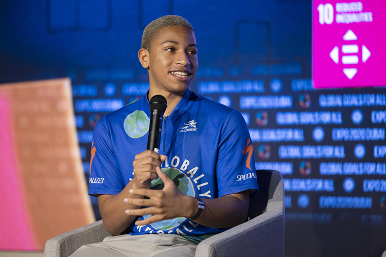 Jerome wears a blue shirt with a globe on it while talking into a microphone.