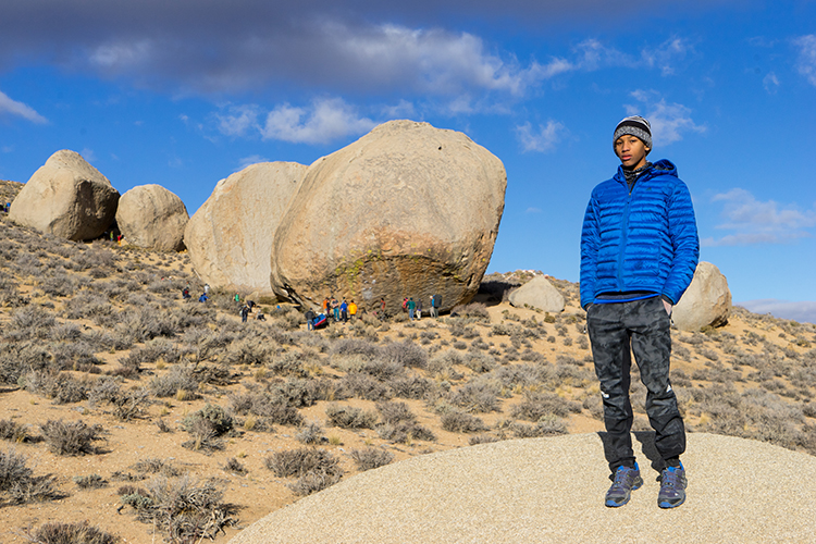 Kai stands in the forefront on a rock with his hands in his pockets and a blue jacket. In the background, we can see people at the base of a massive boulder. 