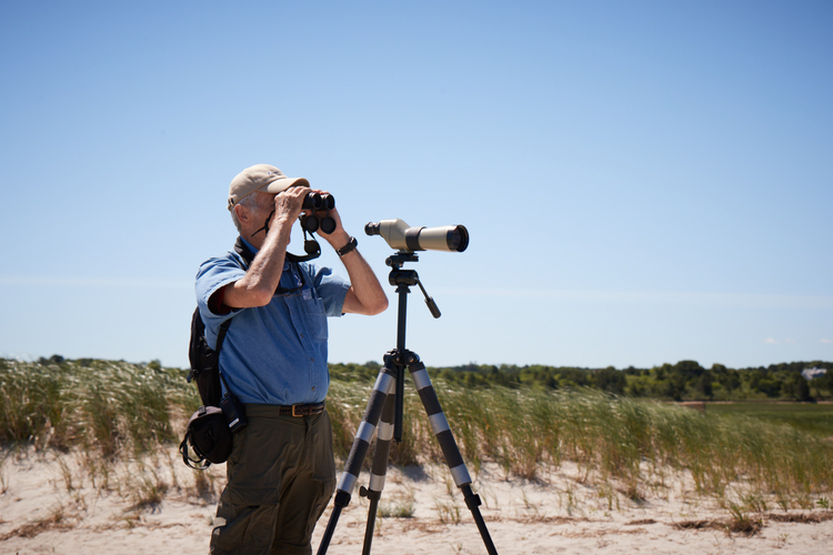 A man standing on sand dunes looking through a pair of binoculars. A scope on a tripod is set up in front of him.