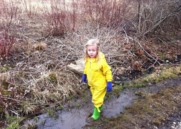 A young blonde girl with green rainboots and blue gloves. She is standing on a muddy trail.