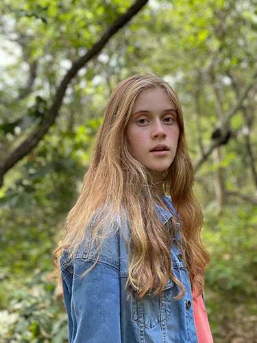 A teenage girl looking at the camera with long, blonde hair and wearing a denim jacket. 