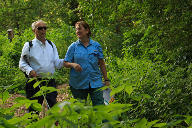 A man in a white button-down shirt with a sight stick and sunglasses, holding one hand onto the elbow of a woman with a blue shirt. They are walking in the woods.